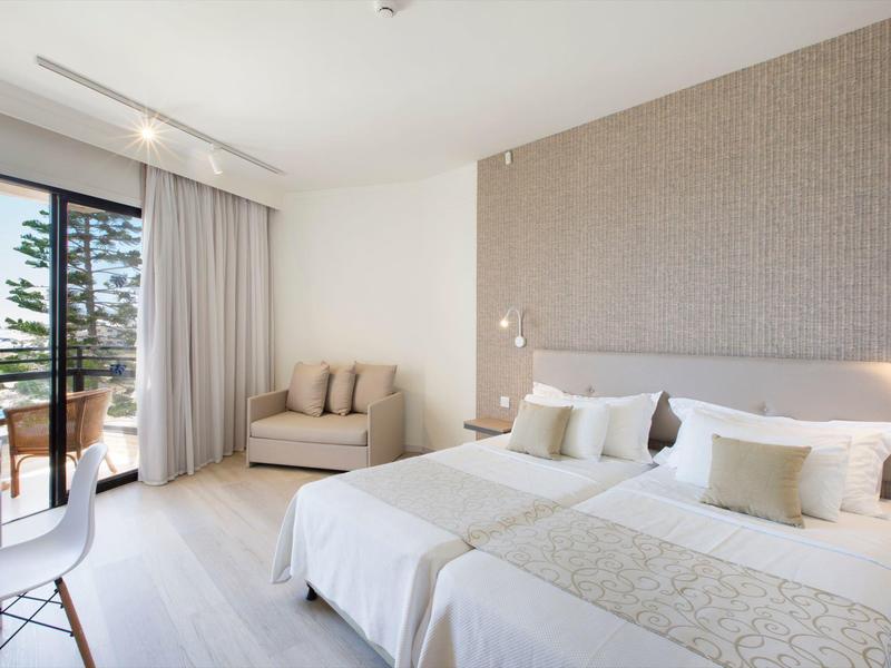 Helles Hotelzimmer mit zwei Einzelbetten, Sitzbereich, Schreibtisch und Balkon mit Meerblick.