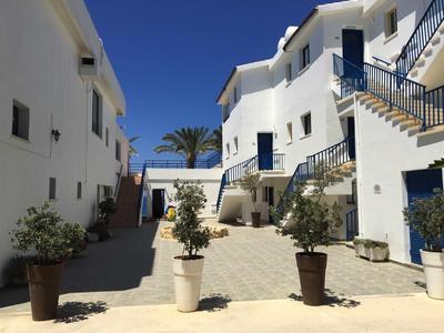 Weiß gestrichene Gebäude mit blauen Türen und Treppen, Pflanzen in Töpfen, sonniger Himmel.