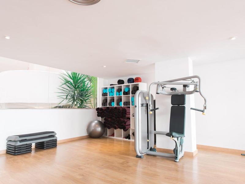 Salle de sport lumineuse avec rack d'haltères, machine d'entraînement, ballons de gym et marches sur sol en bois.