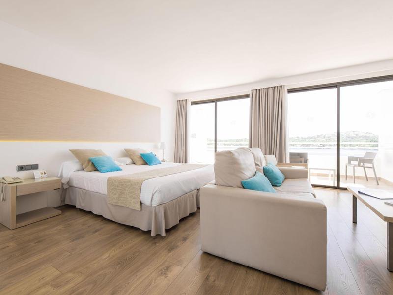 Chambre d'hôtel moderne avec lit double, canapé, sol en bois et grande fenêtre avec balcon.