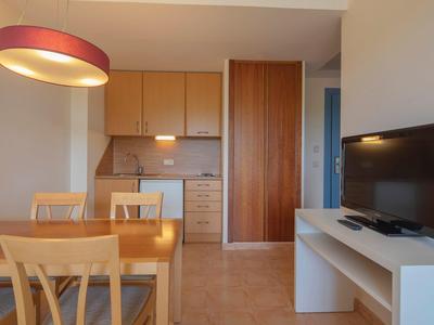 Angolo cottura con armadietti in legno, tavolo da pranzo e sedie, TV su mobile bianco.