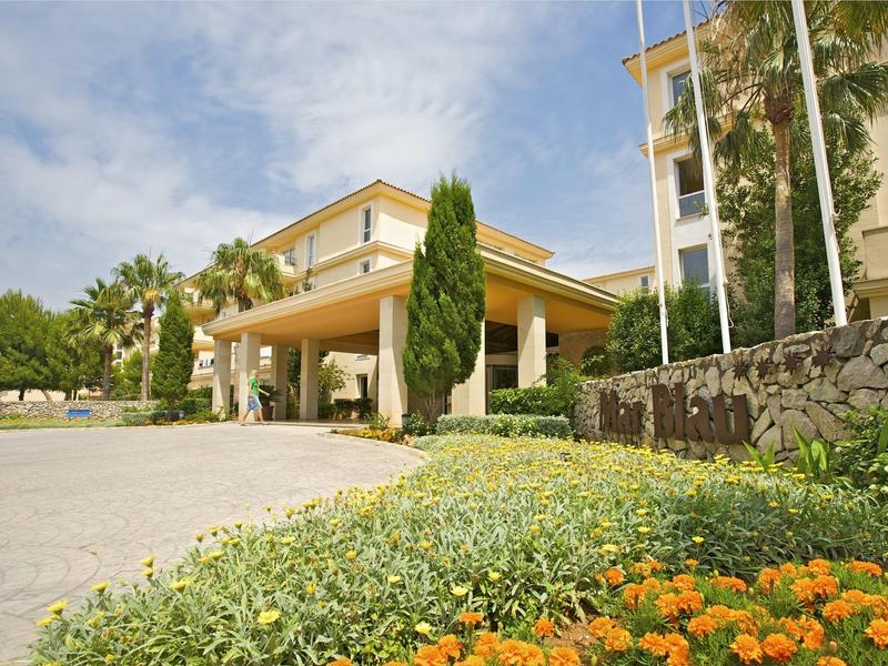 Ingresso dell'hotel con pensilina circondato da cespugli verdi e fiori arancioni sotto un cielo blu.