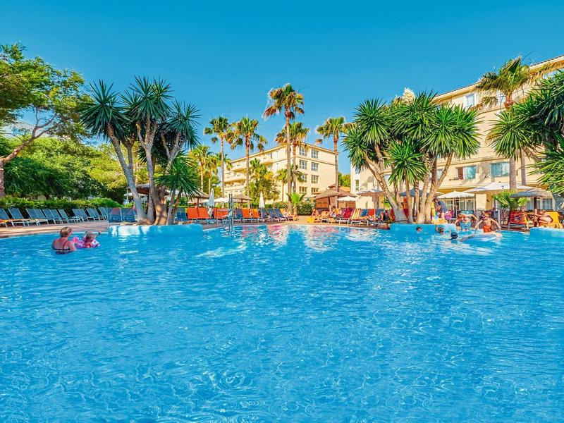 Grande piscina con palme e lettini davanti a un hotel in una giornata di sole