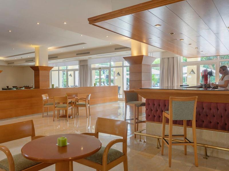 Interno di hotel luminoso con bar, mobili in legno e grandi finestre con vista esterna.