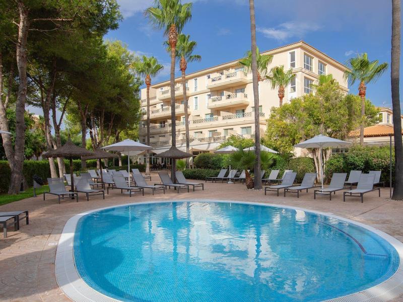 Piscina rotonda con lettini e ombrelloni davanti a un edificio alberghiero e palme.