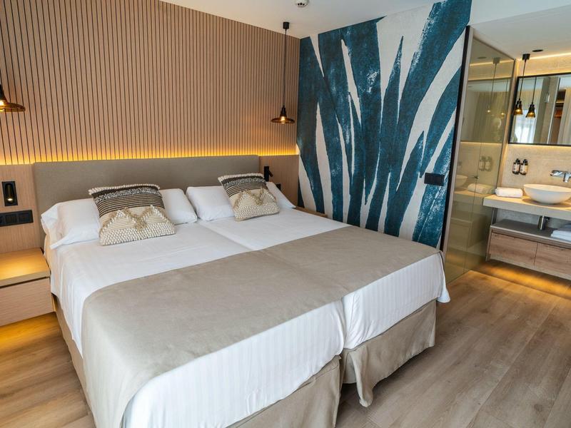 Chambre d'hôtel moderne avec deux lits simples, éclairage chaleureux et salle de bain vitrée.