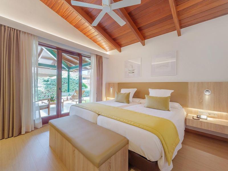 Chambre d'hôtel lumineuse avec deux lits, plafond en bois et accès à un balcon avec vue sur la verdure.