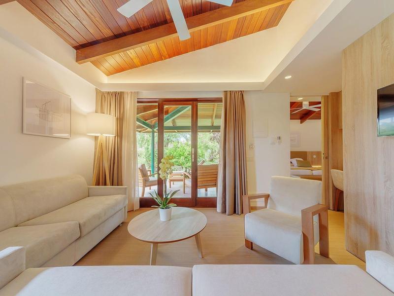 Salon lumineux avec plafond en bois, canapé, fauteuils et accès à une terrasse donnant sur la verdure.