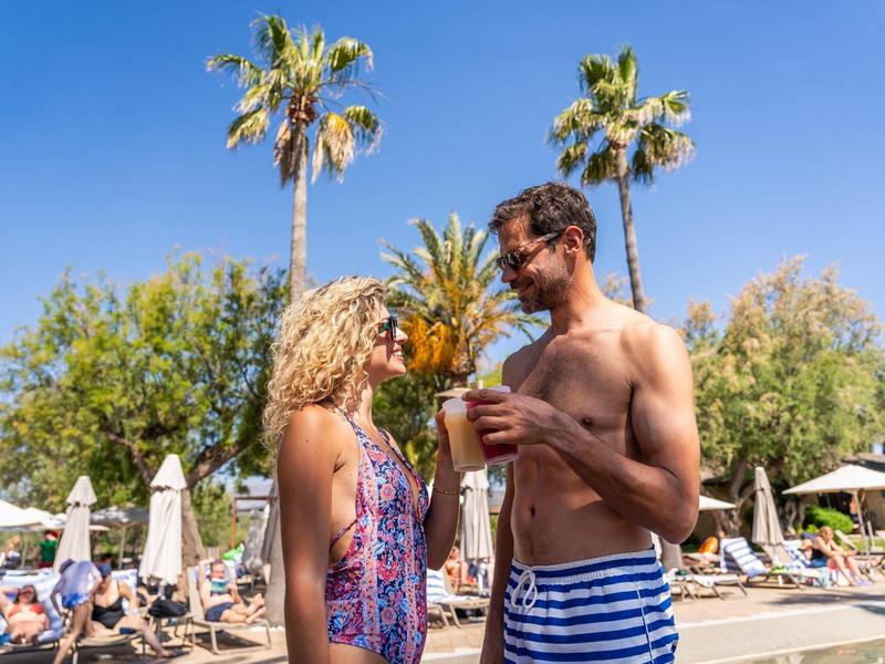 Couple en maillot de bain dégustant des boissons au bord de la piscine avec palmiers et chaises longues.