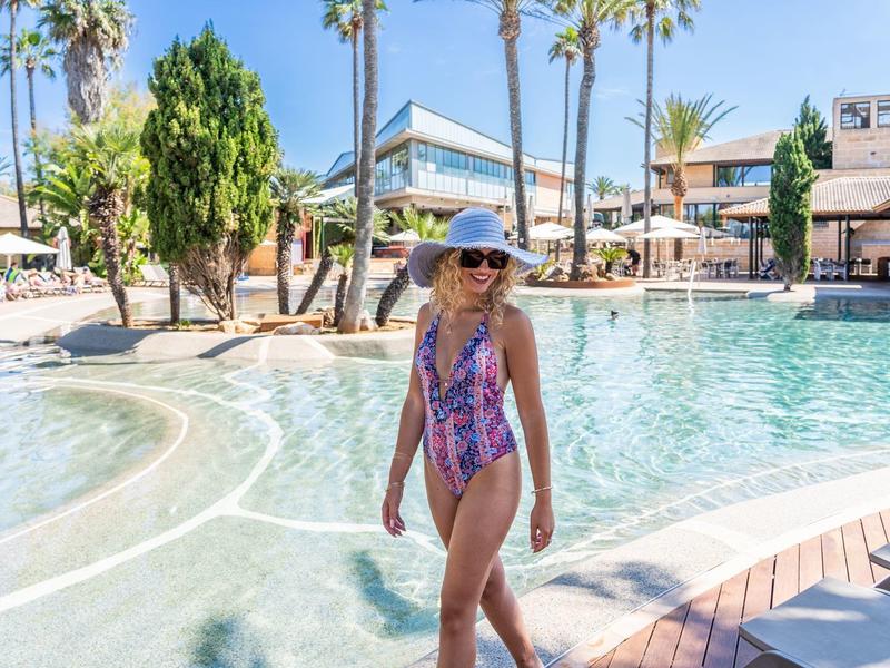 Femme en maillot de bain et chapeau marche le long d'une piscine d'hôtel par une journée ensoleillée.