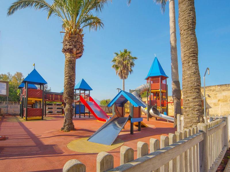 Aire de jeux colorée avec toboggans et structures d'escalade entre des palmiers sous un ciel bleu.