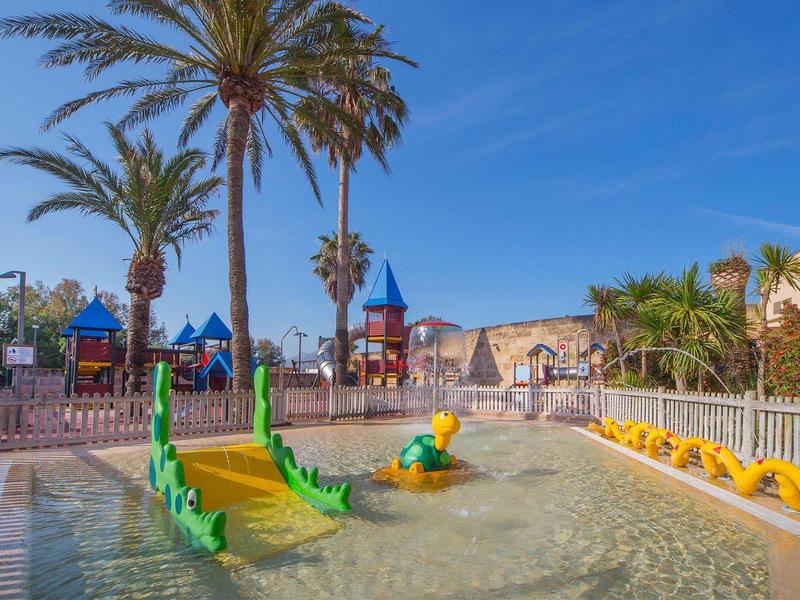 Parc aquatique pour enfants avec fontaines vertes et jaunes près des palmiers et d'une aire de jeux.