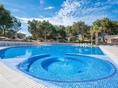 Grande piscina all'aperto con gradini arrotondati, circondata da alberi e lettini sotto un cielo azzurro.