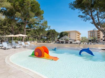 Piscina per bambini con giochi colorati, lettini e edifici dell'hotel sotto un cielo limpido.