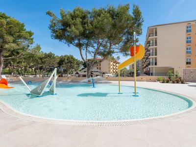 Rundes Planschbecken mit Wasserspielen und bunten Elementen in sonnigem Außenbereich eines Hotels.