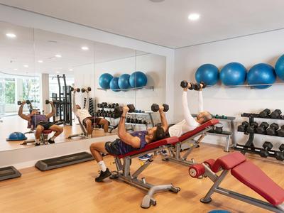 Person trainiert mit Kurzhanteln auf einer roten Bank in einem Fitnessraum mit Spiegeln und blaue Gymnastikbällen.