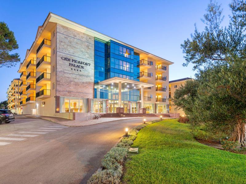 Moderno edificio alberghiero su più piani al tramonto con balconi illuminati e parcheggio.