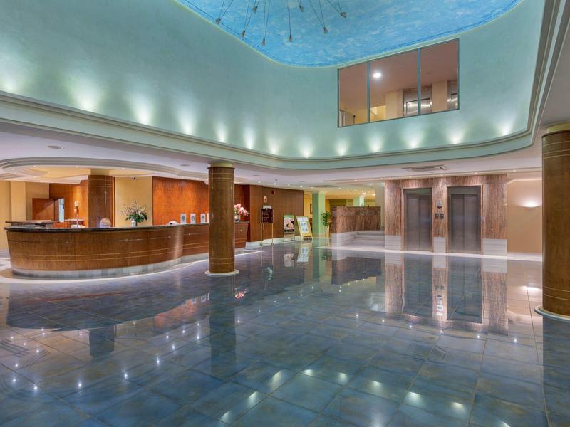 Ampia hall d'hotel con pavimento lucido, banco reception e illuminazione moderna sotto soffitto in vetro.