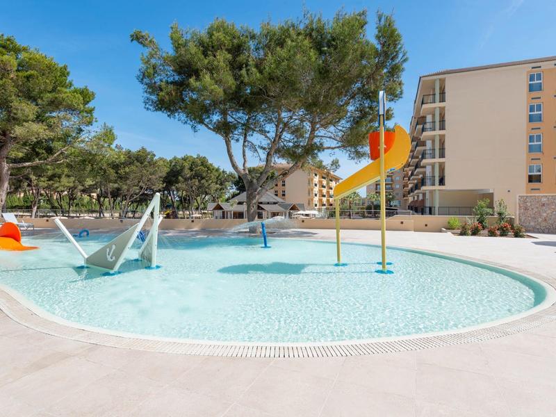 Piscina circolare per bambini con giochi d'acqua circondata da alberi e edifici.