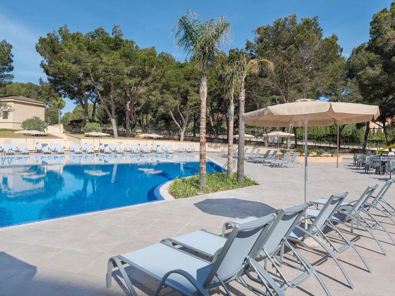 Lettini moderni accanto a una piscina limpida con ombrelloni e alberi sullo sfondo.