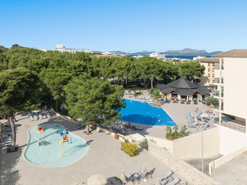 Terrazza dell'hotel con piscina, ombrelloni e area giochi accanto a una zona boschiva.