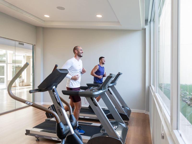 Due persone corrono su tapis roulant in una palestra luminosa con grandi finestre.