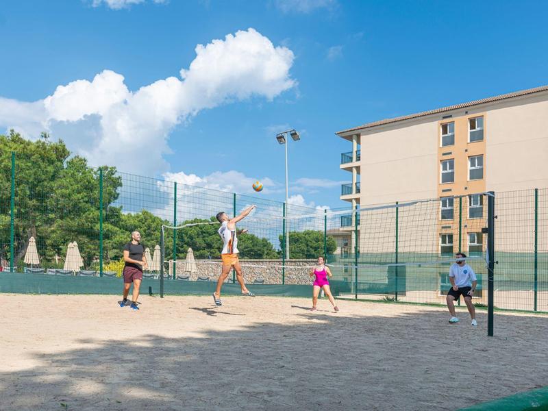 Famiglia che gioca a pallavolo su un campo sabbioso accanto a un edificio dell'hotel in una giornata di sole.