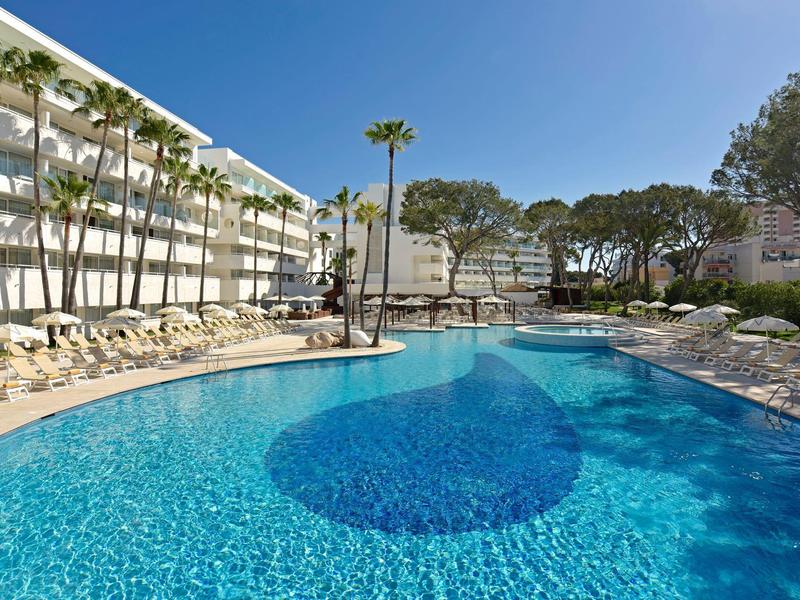 Velký hotelový bazén s čistou modrou vodou, obklopený palmami a lehátky pod jasnou oblohou.