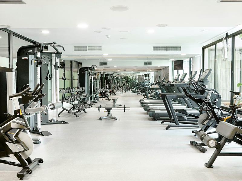 Moderní posilovna s kardio a posilovacími přístroji v světlé hotelové místnosti.