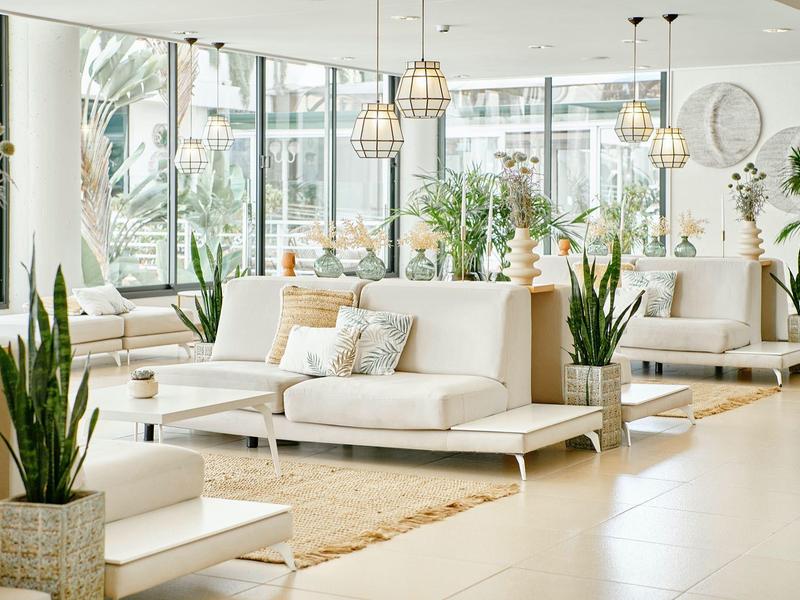 Světlý, moderní hotelový salonek s bílými pohovkami, rostlinami a velkými okny.