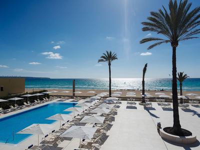 Hotelový bazén s lehátky a výhledem na moře s palmami pod modrou oblohou.