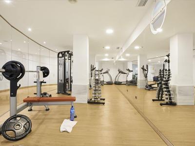 Heller Fitnessraum mit Spiegelwand, Hanteln, Langhantel und Trainingsgeräten.