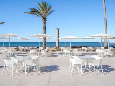 Leerer Außenbereich mit weißen Tischen und Stühlen vor dem Meer unter blauem Himmel.