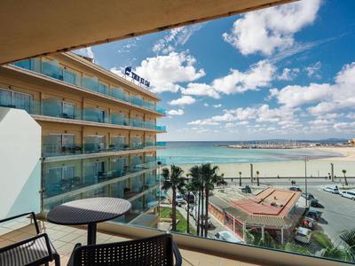 Balkon mit Tisch und Stühlen mit Blick auf Hotel, Strand und Meer bei sonnigem Wetter.
