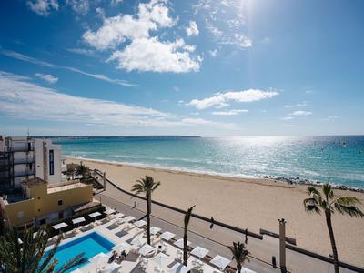Blick auf einen sonnigen Strand mit Palmen, Poolbereich und Hotelterrasse unter blauem Himmel.