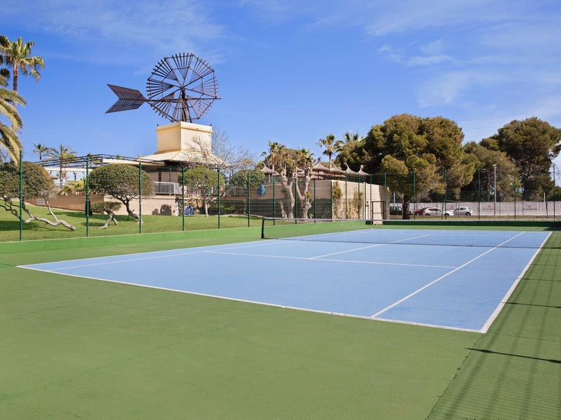 Venkovní tenisový kurt s větrným mlýnem a stromy v pozadí pod modrou oblohou.