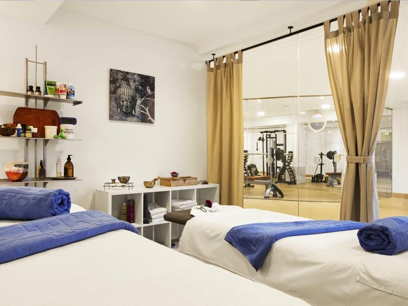 Spa místnost s masážními stoly, modrými ručníky a výhledem do posilovny.