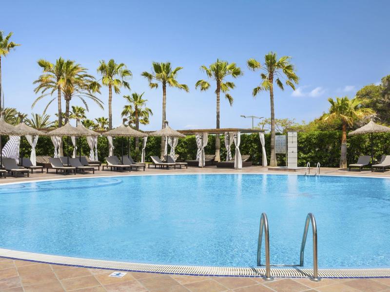 Venkovní hotelový bazén s lehátky a palmami pod modrou oblohou.