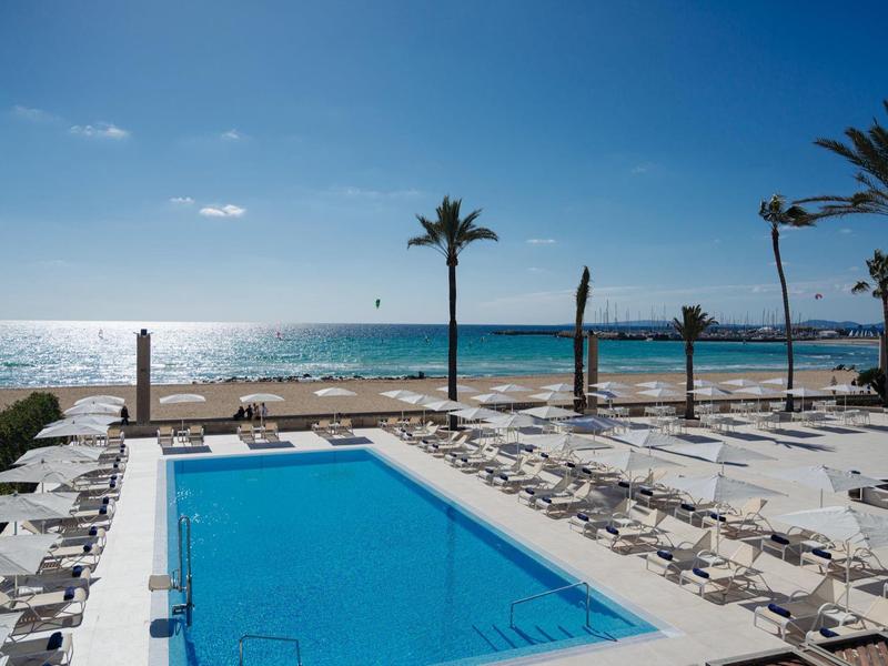 Modrý bazén s výhledem na moře, obklopený lehátky a palmami pod jasnou oblohou.