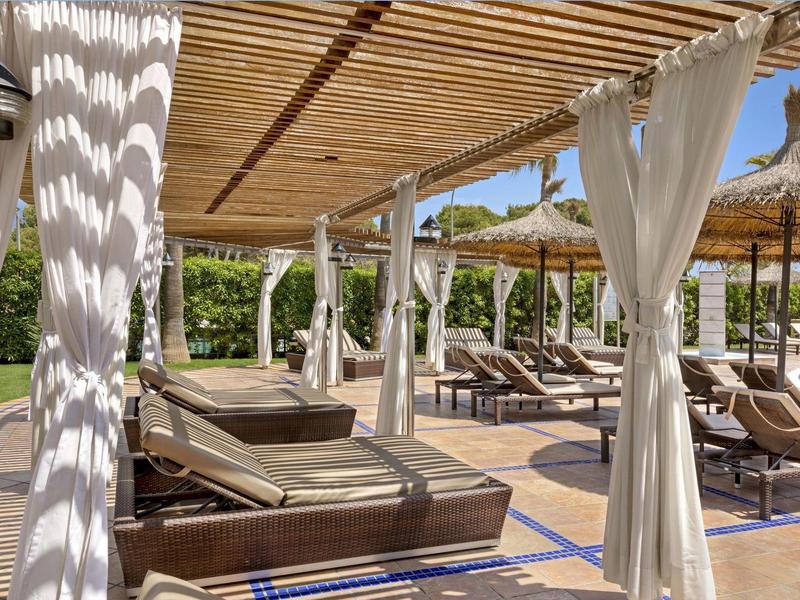 Hotelová terasa s lehátky pod dřevěnou pergolou a bílými záclonami.