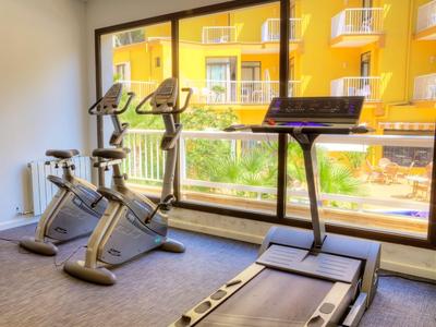 Fitnessraum mit zwei Fahrradergometern und einem Laufband vor großen Fenstern mit Blick auf ein gelbes Gebäude.