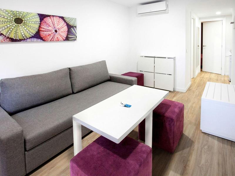 Salon moderne avec canapé gris, table blanche et tabourets colorés.