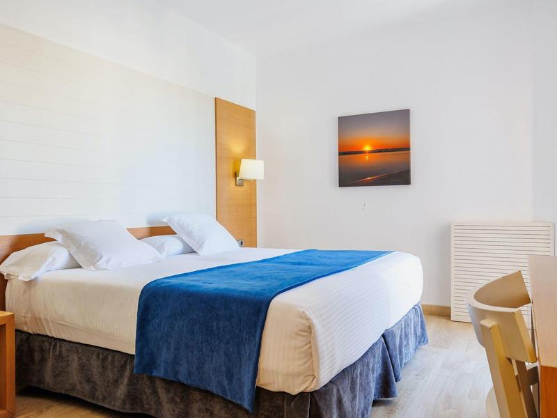 Chambre d'hôtel lumineuse avec un lit double, une couverture bleue et une photo de coucher de soleil au mur