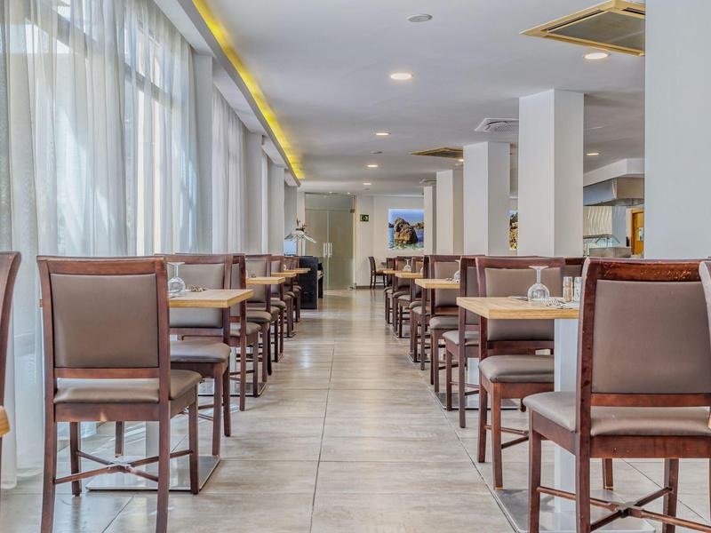 Salle à manger vide avec des chaises et tables marron, éclairée par de grandes fenêtres.
