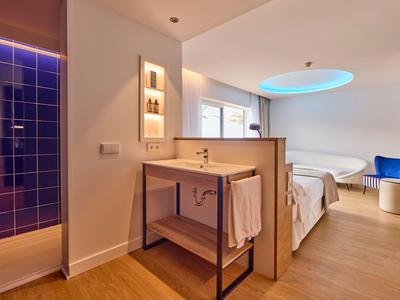 Modern hotel room with wooden floor, double bed, and an illuminated bathroom shower area.