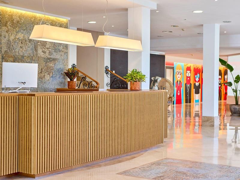 Modern hotel reception area with wooden desk, lamps, and plants in a bright lobby.