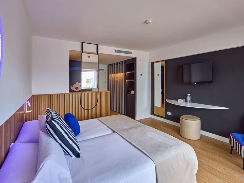 Modern hotel room with large bed, purple lighting, and seating area with chair and ottoman.