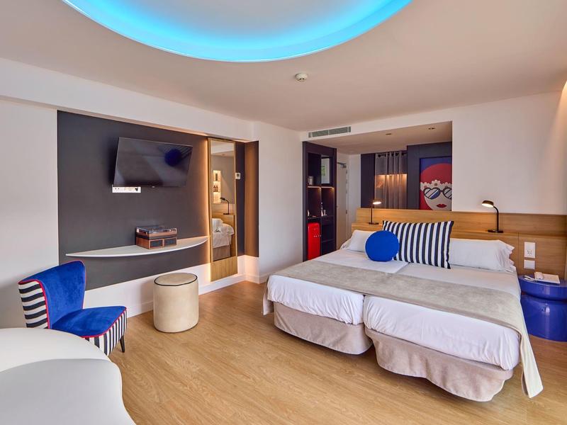 Modern hotel room with twin beds, cozy seating, and soft wooden flooring under blue ambient lighting.