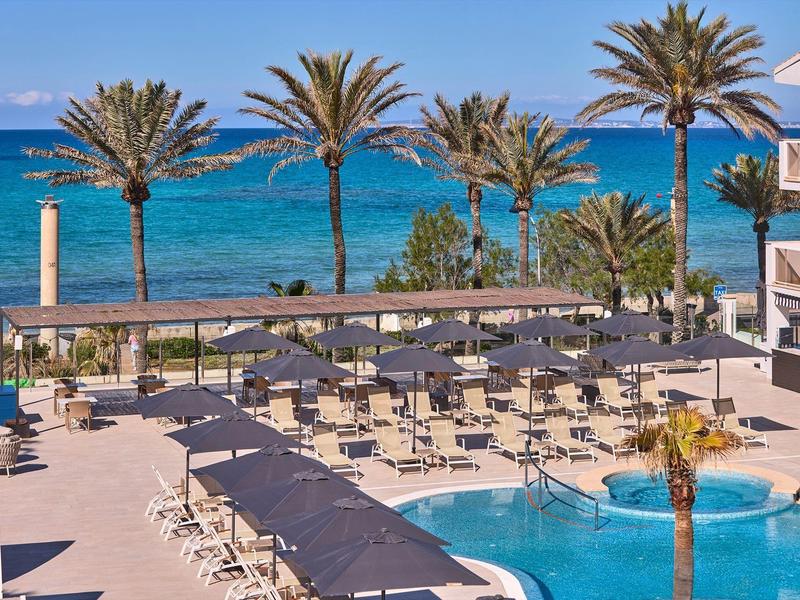 Hotel pool area with lounge chairs, palm trees, and a view of the blue sea under clear sky.