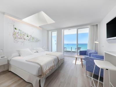 Helles Hotelzimmer mit großem Bett, blauen Sesseln und Meerblick durch bodentiefe Fenster.